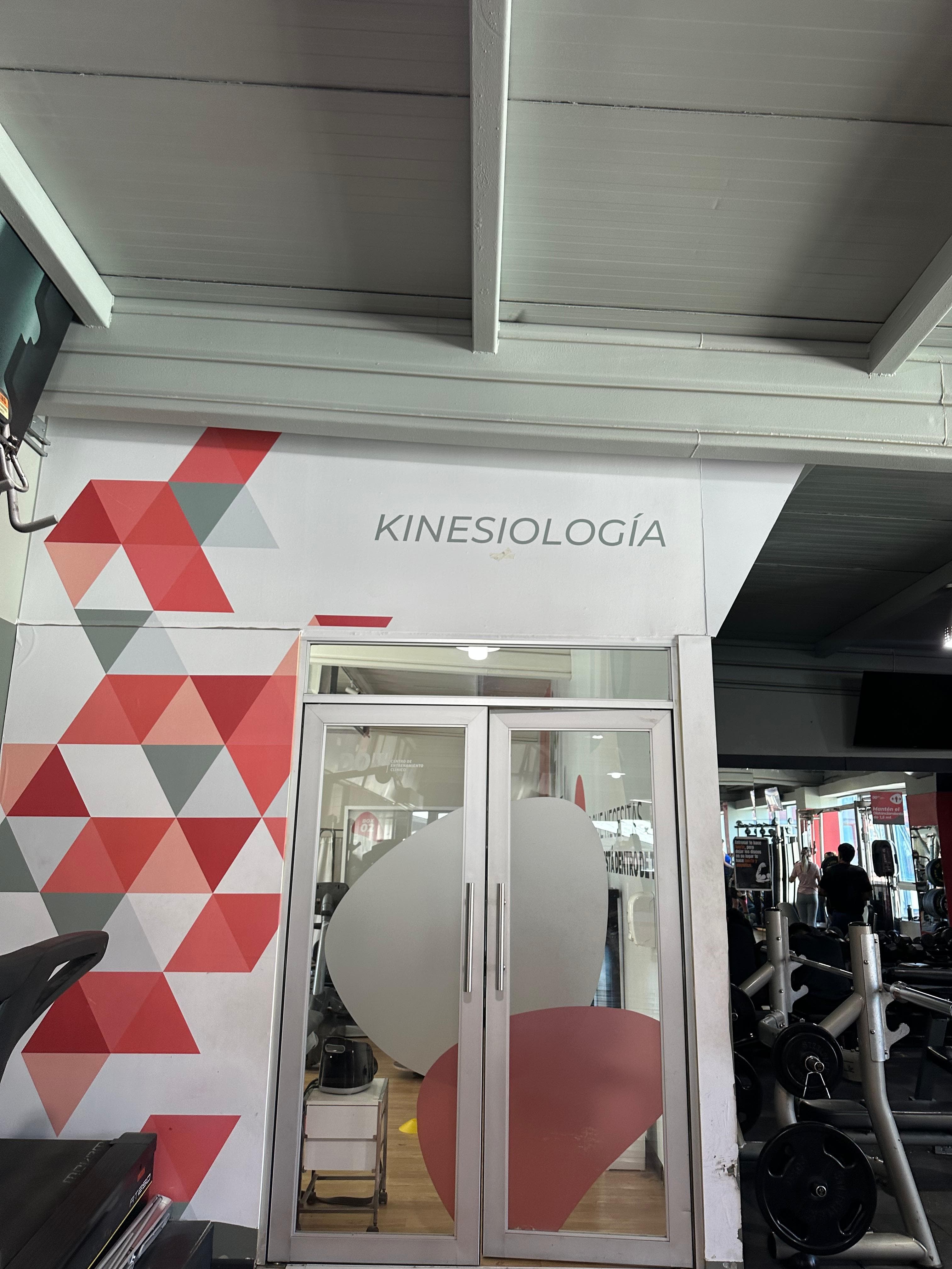 Entrada al box de kinesiología de R3SALUD en Do It Fitness: puertas de vidrio bajo el letrero Kinesiología y un muro con diseño geométrico